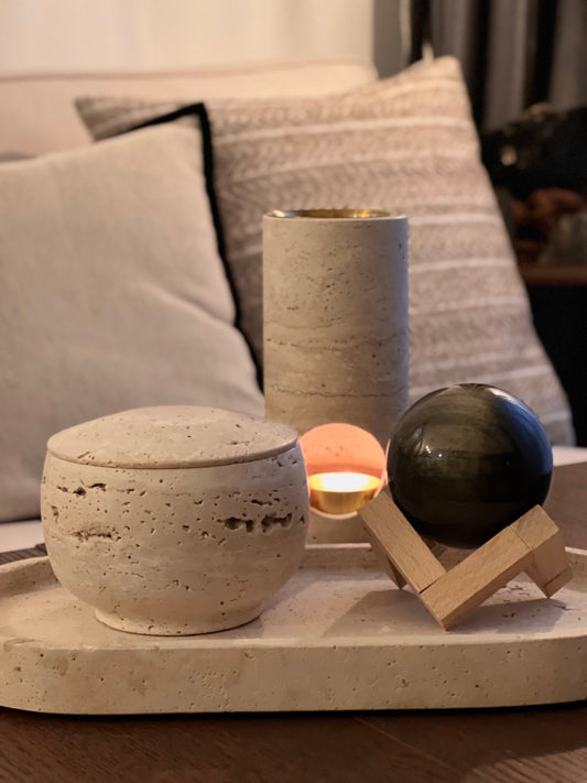 Decorative items including a travertine scented candle, a travertine oil or wax melts burner and a golden obsidian sphere on a wooden surface with pillows in the background.