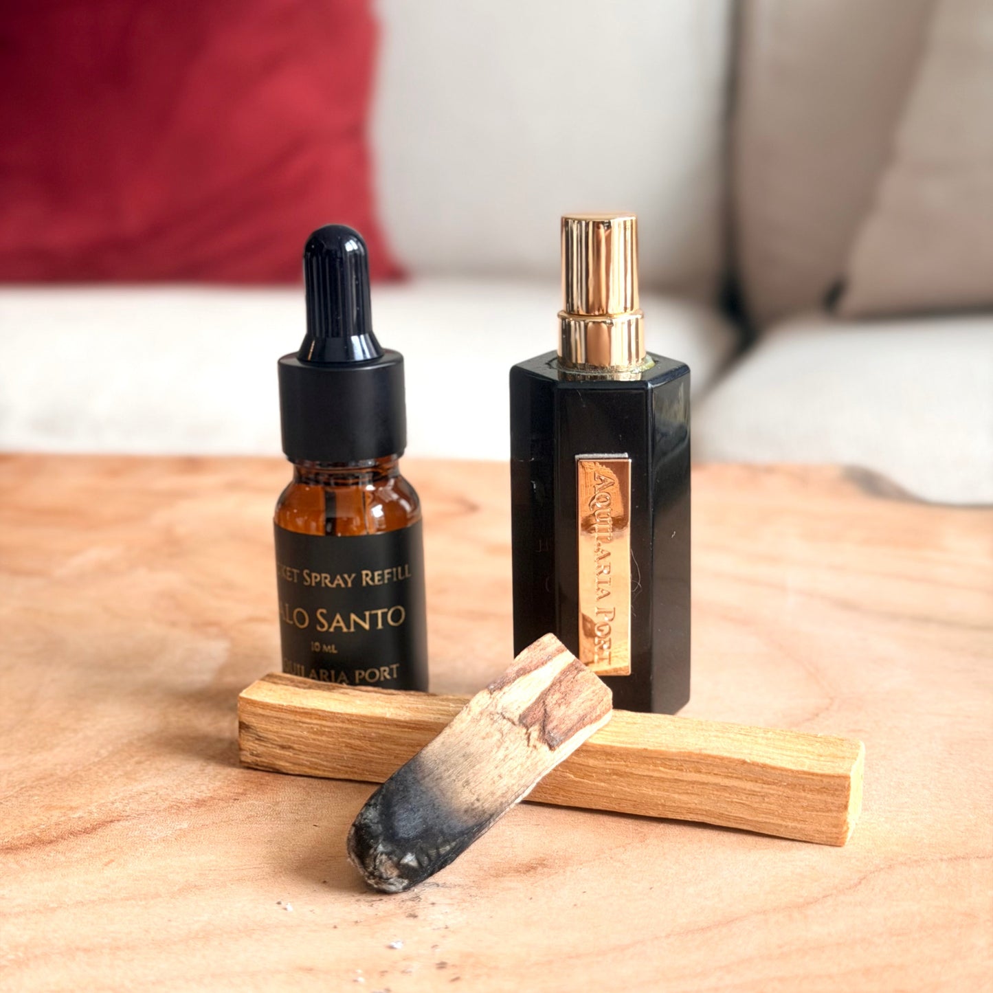 A black obsidian atomiser and a 10ml dropper bottle of "Palo Santo" Pocket Spray refill Aquilaria Port standing on a light wood surface with a blurred sofa and red pillow in the background. 