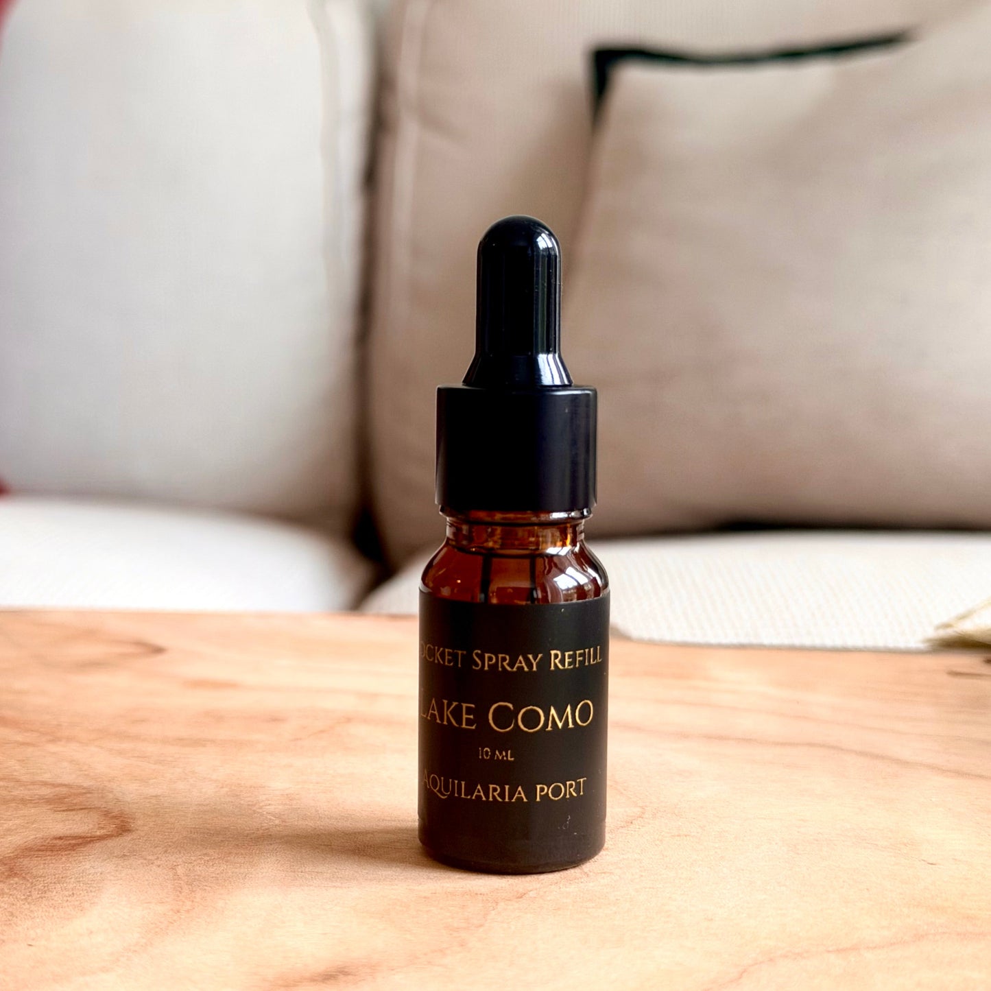 A small amber glass dropper bottle labeled 'LAKE COMO' and 'AQUILARIA PORT' sits on a light wood tabletop with a blurred sofa in the background.