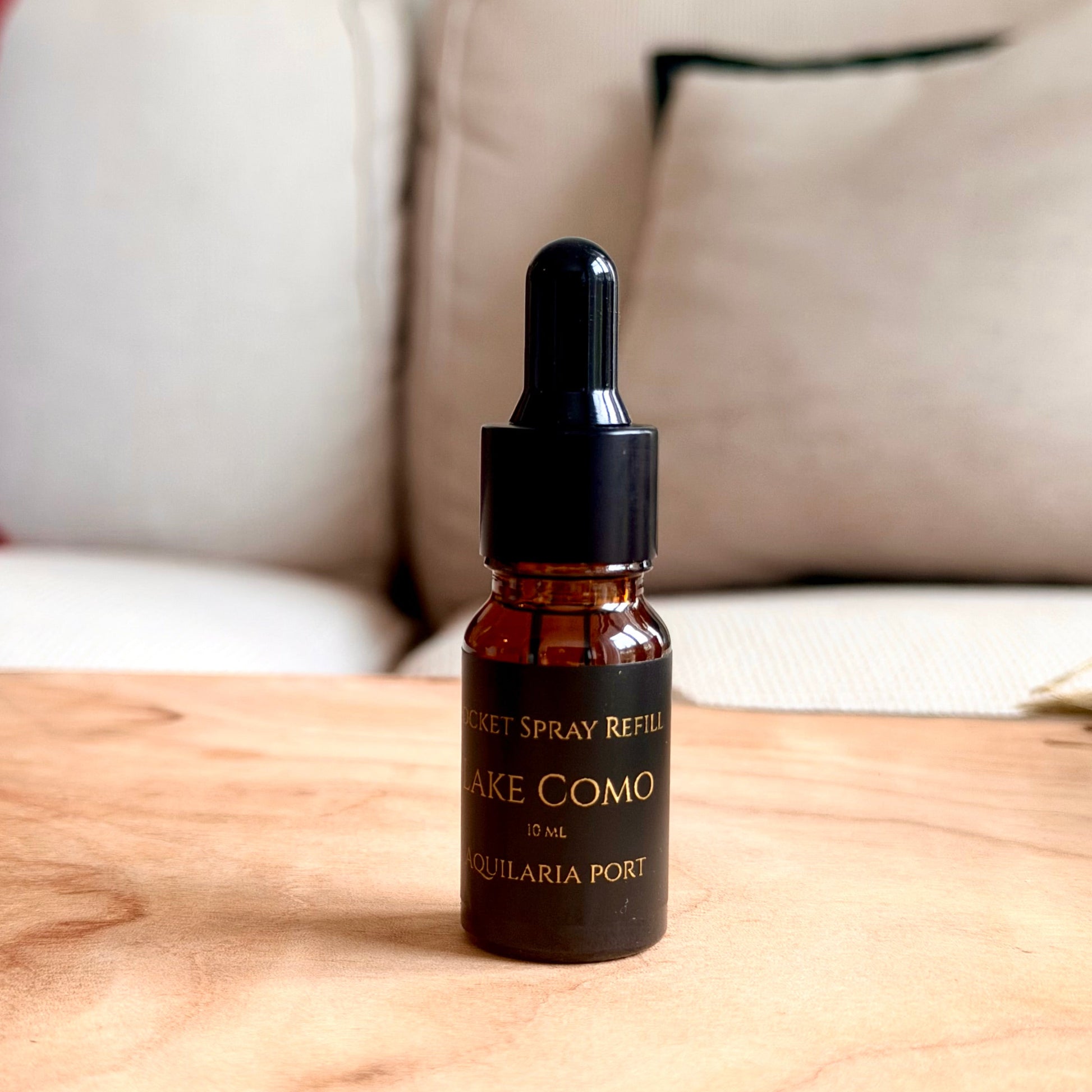 A small amber glass dropper bottle labeled 'LAKE COMO' and 'AQUILARIA PORT' sits on a light wood tabletop with a blurred sofa in the background.