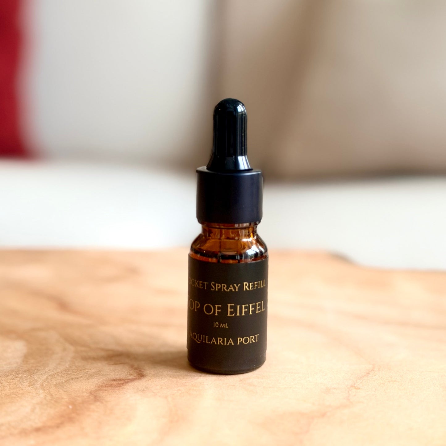 A small amber glass dropper bottle with a black cap stands on a light wood surface in a softly blurred indoor setting with a sofa and pillows in the background.