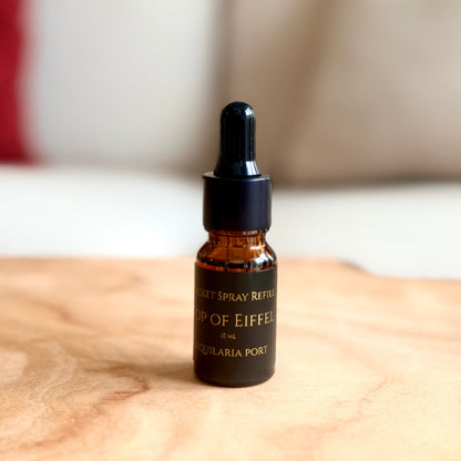 A small amber glass dropper bottle with a black cap stands on a light wood surface in a softly blurred indoor setting with a sofa and pillows in the background.