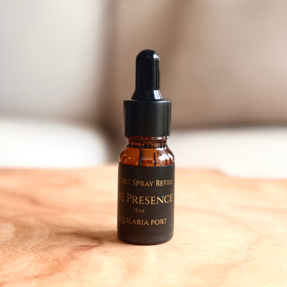 A small amber glass dropper bottle labeled 'POCKET SPRAY REFILL' and 'THE PRESENCE by AQUILARIA PORT' sits on a light wood surface with a softly blurred indoor background.