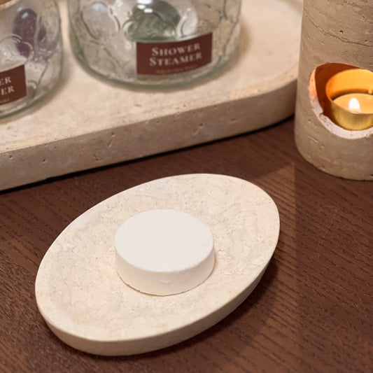 White stone-like soap dish with a bar of soap on a wooden surface, with a candle in the background.