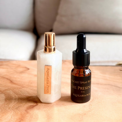 A white quartz atomiser and a 10ml dropper bottle of "The Presence" Pocket Spray refill Aquilaria Port standing on a light wood surface with a blurred sofa and red pillow in the background. 
