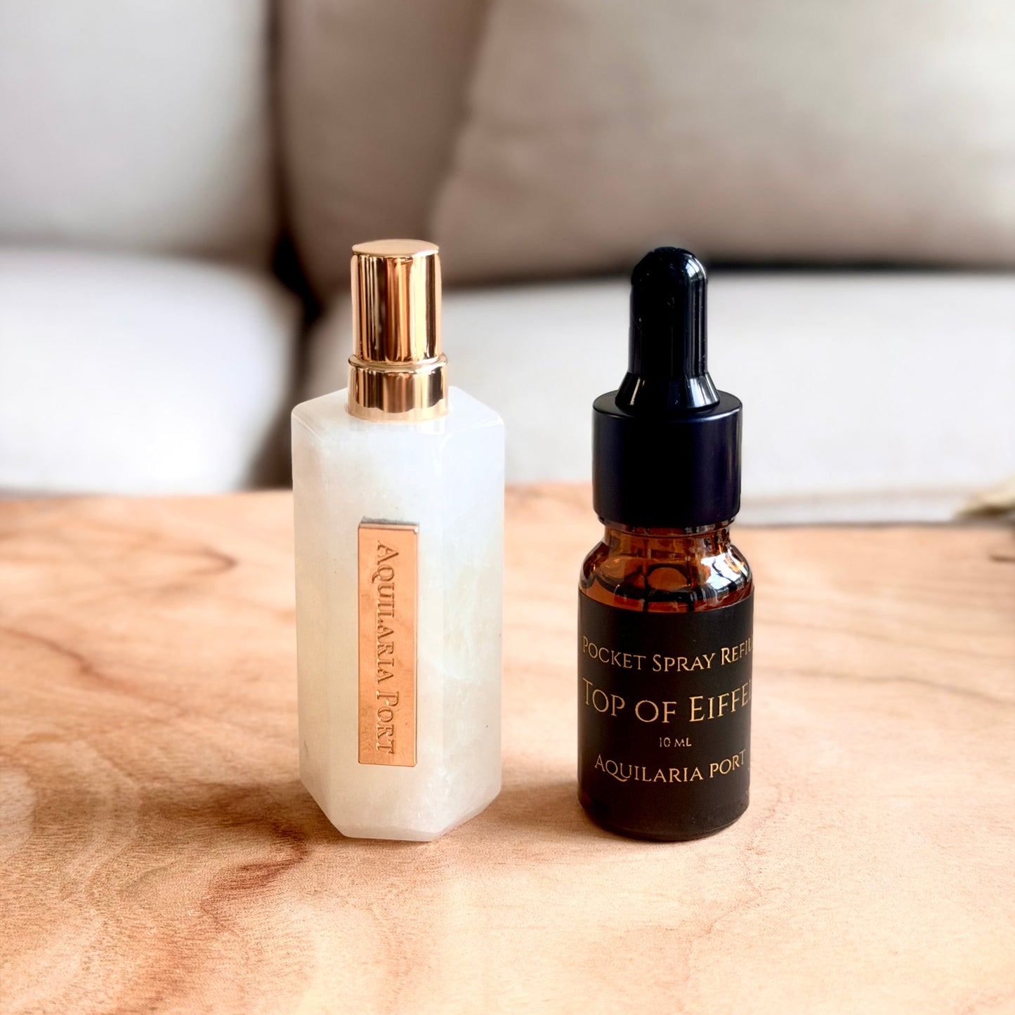 A white quartz atomiser and a 10ml dropper bottle of "Top of Eiffel" Pocket Spray refill Aquilaria Port standing on a light wood surface with a blurred sofa and red pillow in the background. 