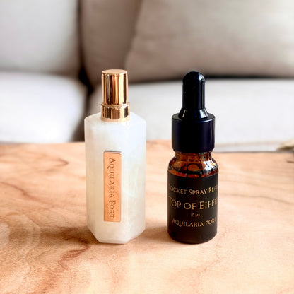 A white quartz atomiser and a 10ml dropper bottle of "Top of Eiffel" Pocket Spray refill Aquilaria Port standing on a light wood surface with a blurred sofa and red pillow in the background. 