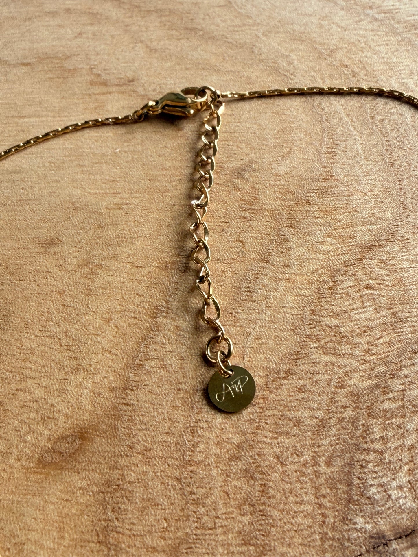 Gold necklace extension with the logo AqP with a round pendant on a wood textured background